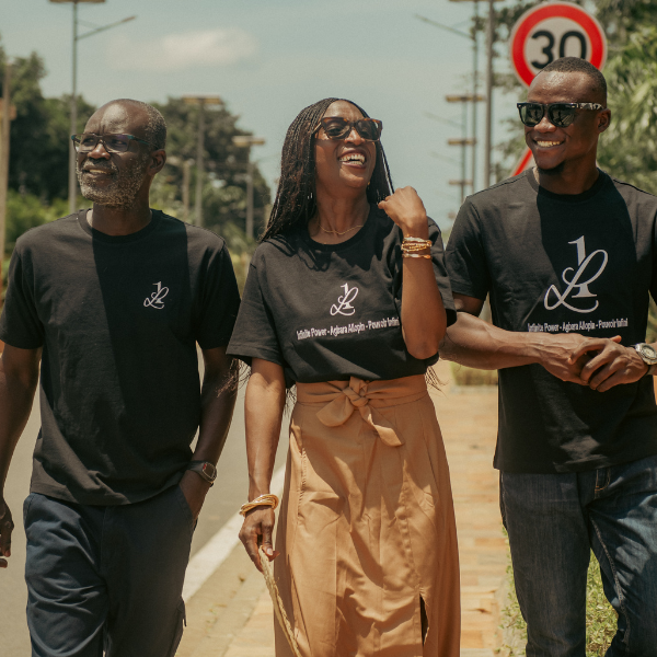 Trois personnes souriantes portant des t-shirts noirs 1Look by Adé avec le slogan "Infinite Power – Agbara Alopin", marchant dans une rue au Bénin, incarnant la vision de la marque entre héritage, style et singularité.